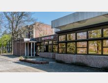 Ein flaches Gebäude mit großen Fensterfronten und einer Eingangstür unter einem kleinen Vordach, beschriftet mit "Ritschi's Treff". Im Hintergrund sind Bäume und ein mehrstöckiger Wohnblock zu sehen.