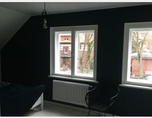 Ein Zimmer mit dunkel gestrichenen Wänden, drei Fenstern, die Tageslicht hereinlassen, und Blick auf eine Außenumgebung mit Bäumen und Gebäuden. Im Innenraum sind ein Heizkörper, ein schwarzer Stuhl sowie ein Bett sichtbar.