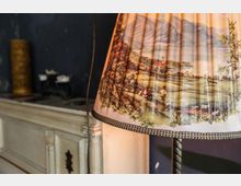 Nahaufnahme einer Stehlampe mit einem Schirm, der eine ländliche Landschaft mit Bergen, Bauernhofgebäuden, Menschen und Tieren darstellt. Im Hintergrund ist ein antiker weißer Kamin und eine dunkle Wand mit dekorativen Elementen sichtbar.