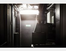 Technikraum mit schwarzer Tür, auf der ein Schild mit der Aufschrift "Notausgang" angebracht ist. Sichtbare Rohrleitungen und technische Geräte dominieren den engen Raum, der von grellem Licht beleuchtet wird.