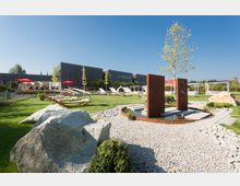 Weitläufige Gartenlandschaft mit gepflegtem Rasen, modernen Liegestühlen, roten Sonnenschirmen und einer Wasserinstallation aus Metallplatten umgeben von Kies. Im Hintergrund befinden sich moderne Gebäude mit dunkler Holzverkleidung.