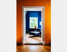 Blick durch einen weißen Türrahmen aus einem Raum mit orangefarbenen Wänden in ein Wohnzimmer mit dunkelblauer Wand, großem Sprossenfenster und blauem Raffrollo. Vor dem Fenster stehen ein dunkler Sessel mit Ottomane, ein kleiner Tisch mit Vase sowie ein gemusterter Teppich auf Holzboden.