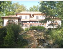 Ein zweistöckiges Gebäude mit heller Fassade, roten Dachziegeln und großen Fenstern, umgeben von dichter Vegetation. Im Vordergrund führt ein kleiner Steingarten mit einem Bachlauf durch die Landschaft.