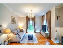 Wohnzimmer mit eleganter Einrichtung: ein cremefarbener Sofa mit Kissen, ein zentral platzierter Couchtisch und ein großer Flachbildfernseher auf einer niedrigen Konsole. Der Raum verfügt über hölzernes Fischgrätenparkett, einen Kronleuchter und große Fenster mit schweren Vorhängen, die auf einen Garten blicken.