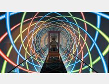 Ein schmaler, geschlossener Gang mit Glasgeländern, der von bunten, sich überlappenden Neonlichtringen in Blau, Rot, Grün und Gelb umgeben ist, bildet eine futuristische, spiralförmige Lichtinstallation. Der Gang endet in einem rechteckigen Rahmen und spiegelt das Licht auf der glänzenden Bodenfläche wider.
