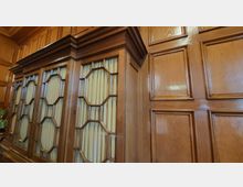 Holzgetäfelter Raum mit einer Wand aus glänzendem Holz und einem eingebauten Schrank. Der Schrank hat geometrisch unterteilte Glaselemente mit dahinterliegenden gelblichen Vorhängen.