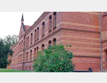 Backstein-Gebäude im historistischen Stil mit mehreren Etagen und rundbogigen Fenstern, verziert mit gelben Zierbändern; im Vordergrund ein Busch und eine Grünfläche, dahinter Bäume.