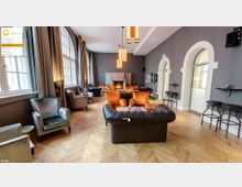 Gemütlicher Loungebereich mit modernem Kamin, Leder- und Samtsesseln in warmen Farben, Parkettboden und großen Rundbogenfenstern mit Vorhängen in einem eleganten, grauen Innenraum.