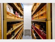 Ein schmaler Lagerraum mit hohen Regalreihen, die mit gelben und roten Plastikboxen gefüllt sind. Eine weiße Backsteinwand mit einem Heizkörper bildet den Abschluss des Raumes.