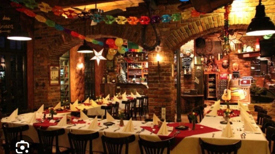 Gemütlicher Restaurantinnenraum mit Backsteinwänden und Rundbögen, langen gedeckten Tischen mit weißen Tischdecken und gefalteten Servietten. Bunte Girlanden, Ballons und Hängelampen schmücken den Raum, im Hintergrund ist ein kleiner Barbereich mit Regalen zu sehen.