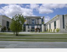 Moderne Regierungsgebäude mit einer breiten Glasfassade und Betonarchitektur, umgeben von gepflegten Rasenflächen, Bäumen und einem großen Metallkunstwerk im Vordergrund. Der Himmel im Hintergrund ist bewölkt mit vereinzeltem Sonnenlicht.