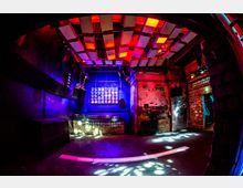 Innenansicht eines Nachtclubs mit Backsteinwänden, einer beleuchteten Tanzfläche und einer Decke mit modernen, hängenden Paneelen in Rot- und Violetttönen. Eine Wand ist mit einer großen LED-Lichtinstallation versehen, die den Raum farbig erhellt.