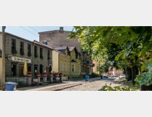 Straßenszene mit historischen Gebäuden, darunter ein Gasthaus mit der Aufschrift „Zu den drei Linden“, und gelben sowie beigegetönten Fassaden. Kopfsteinpflaster, große Bäume auf der rechten Seite und parkende Autos säumen die ruhige Umgebung.