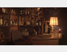 Gemütliche Hausbar mit Holztheke, umgeben von Regalen, die mit verschiedenen Alkoholflaschen und Gläsern gefüllt sind. Eine Tischlampe und eine brennende Kerze sorgen für warmes, stimmungsvolles Licht.