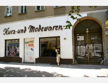 Fassade eines älteren Gebäudes mit weißem Anstrich und der Aufschrift „Kurz- und Modewaren“ über zwei großen Schaufenstern. Rechts daneben eine hölzerne Eingangstür mit Graffiti und einem Warnschild, davor steht eine Person mit Rucksack, die in ein Schaufenster schaut.