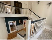 Treppenhaus in einem Wohnhaus mit weißem Holzgeländer und dunkelblauer Wand. Das Geländer führt zu einem oberen Stockwerk, im unteren Bereich sind mehrere Räume mit Holzböden sowie ein goldener Spiegel und eine verzierte Heizung sichtbar.