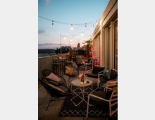 Eine gemütlich gestaltete Dachterrasse bei Dämmerung mit stilvollen Sitzmöbeln, Teppichen und kleinen Tischen, dekoriert mit Kissen, Kerzen und Blumen. Lichterketten spannen sich über den Außenbereich, und im Hintergrund sind Palmen sowie ein städtisches Panorama sichtbar.