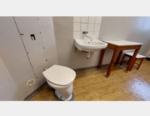 Ein schlichter Raum mit Holzboden, einer Toilette, einem kleinen Waschbecken mit Wandfliesen und einem Holztisch mit zwei Hockern. Die Wände sind weiß gestrichen, und die Einrichtung wirkt funktional.