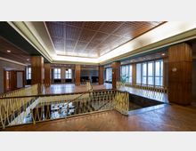 Offener Innenraum eines Gebäudes im Retro-Stil mit Holzverkleidungen, großen Fenstern, einem Geländer im oberen Stockwerk und einem hölzernen Parkettboden. Die Decke ist mit geometrischen Mustern verziert, und Tageslicht fällt durch die Fenster.