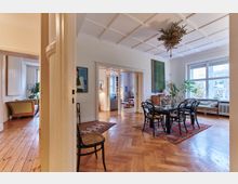 Großzügiger Wohnbereich mit Holzboden, einem Esstisch mit Stühlen auf einem bunten Teppich und großen Fenstern, die viel Tageslicht hereinlassen. Der Raum ist mit klassischen Möbeln, Pflanzen und Kunstwerken an den Wänden dekoriert; im Hintergrund sind angrenzende Zimmer sichtbar.