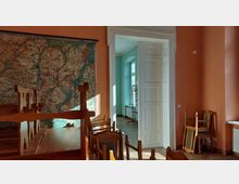 Ein Raum mit orangefarbenen Wänden und einer großen Wandkarte, aufgestellt auf einem Holzständer. Im Vordergrund stehen umgedrehte Holzstühle auf Tischen, während eine geöffnete weiße Tür den Blick in einen angrenzenden, mintfarben gestrichenen Raum mit Fenstern und einem Heizkörper freigibt.