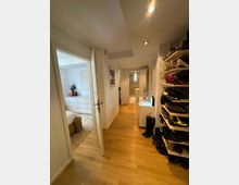 Flur einer Wohnung mit Holzboden, beleuchtet durch deckenintegrierte Spots. Links führt eine Tür in ein Schlafzimmer mit heller Einrichtung, rechts steht ein offenes Regal voller Schuhe, und geradeaus ist ein Badezimmer erkennbar.