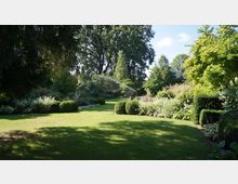 Weitläufiger Garten mit gepflegtem Rasen, umgeben von dichten Sträuchern, Blumenbeeten und Hecken; im Hintergrund stehen große Bäume, und ein Wassersprinkler bewässert die Grünfläche. Die Szenerie ist von Sonnenlicht durchflutet, mit vereinzeltem Schatten von den Bäumen.