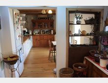 Wohnküche mit rustikalem Holzmobiliar, Holzboden und sichtbaren Holzbalken an der Decke. Auf der linken Seite steht ein Kühlschrank, daneben eine Arbeitsfläche mit Küchengeräten, während rechts ein Durchbruch mit Regal Pflanzen und Dekoration zeigt.