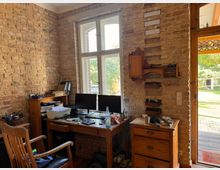 Arbeitszimmer mit rustikaler Backsteinwand, alten Holzmöbeln und moderner Computerausstattung. Ein großes Fenster und eine Glastür lassen Tageslicht herein, mit Blick auf einen Garten im Hintergrund.