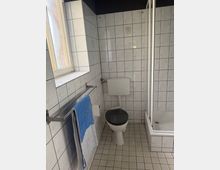 Kleines Badezimmer mit weißen, quadratischen Wand- und Bodenfliesen, Toilette mit Spülkasten an der Wand und schwarzem WC-Sitz. Links ein Milchglasfenster und ein Handtuchhalter, rechts eine Duschwanne mit Glasabtrennung.