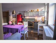 Helles Wohnzimmer-Küchen-Ensemble mit Holzboden, einer roten Kühlschranktür, modernen Geräten und einem Esstisch mit lila Tischdecke. Die Küche hat eine Arbeitsfläche mit Hängeschränken aus mattem Glas, während links ein heller Essbereich mit Stühlen zu sehen ist.