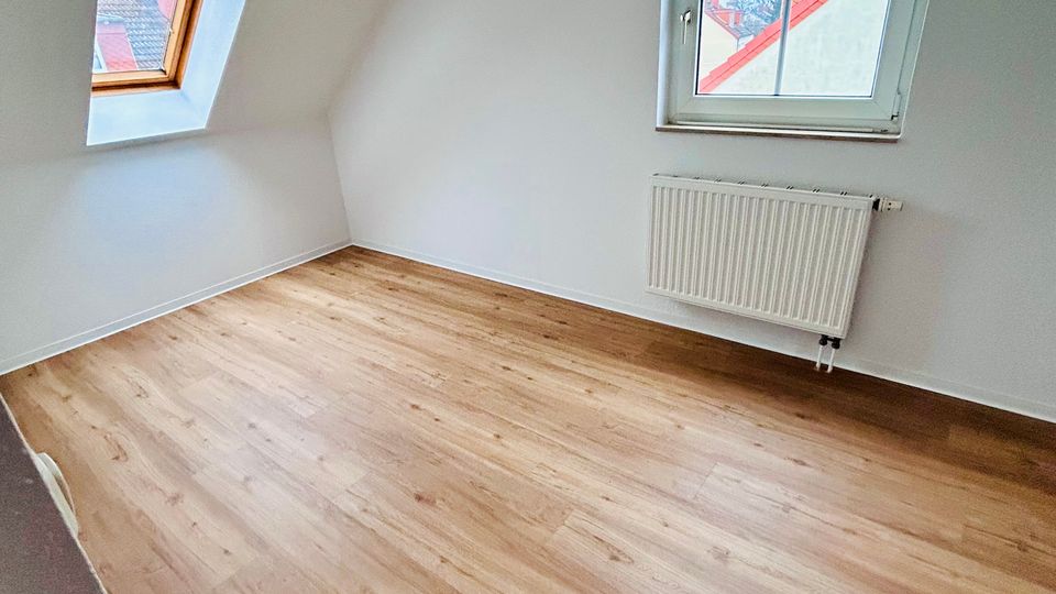 Leerer Dachgeschossraum mit weiß gestrichenen Wänden und schräger Decke, hellen Holzfußboden sowie zwei Fenstern (ein Dachfenster und ein senkrechtes Fenster). Unter dem rechten Fenster befindet sich ein Heizkörper.