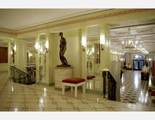 Eleganter Innenraum eines historischen Gebäudes mit Marmorfliesen, Wandleuchten und einem zentralen bronzenen Statuenelement. Im Hintergrund befinden sich große Spiegel, eine dekorative Decke mit Kronleuchtern und ein geometrisches Schachbrettmuster auf dem Boden.