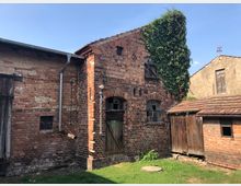 Backsteinbauernhof mit verwitterten Fassaden und einem überwucherten Efeubewuchs an einer Wand. Das Gebäude hat kleine Fenster, eine alte Holztür und ein Schrägdach, teils mit Ziegeln bedeckt; daneben steht ein kleiner Anbau aus Holz.