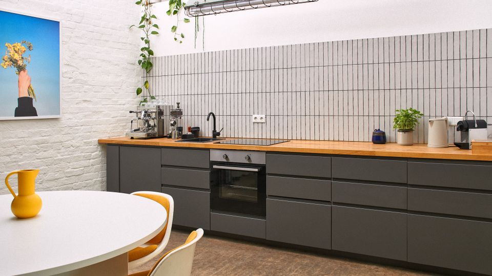 Moderne Küchenzeile mit grauen Unterschränken, Holzarbeitsplatte und vertikal gefliester Rückwand, mit Spüle, Backofen sowie Kaffeemaschine und Wasserkocher. Links stehen ein runder weißer Tisch mit Stühlen und eine gelbe Kanne; an der weißen Ziegelwand hängt ein Bild, darüber eine Hängeleuchte und hängende Pflanzen.