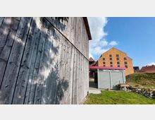 Holzverschaltes Gebäude im Vordergrund mit grober, verwitterter Oberfläche. Im Hintergrund ist ein großes, gelbes Backsteingebäude mit mehreren kleinen Fenstern zu sehen, daneben ein kleines modernes Gebäude mit Metallverkleidung und rotem Dach. Im Vordergrund Wiese und gepflasterte Wege, die Szene wird von blauem Himmel ergänzt.