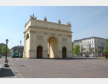 Eine historische Toranlage mit hellem Sandstein und dekorativen Verzierungen, darunter Reliefs und Figuren auf der Spitze, steht auf einem gepflasterten Platz. Im Hintergrund sind Wohnhäuser im klassischen Stil sowie Bäume sichtbar.