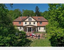 Zweistöckiges Gebäude mit heller Fassade und einem roten Ziegeldach, umgeben von dichter grüner Vegetation. Vor dem Haus befindet sich eine Terrasse, auf der mehrere Menschen an Sitzgruppen unter freiem Himmel zusammensitzen.