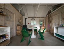 Rustikaler Innenraum mit unverputzten Steinwänden, offenem Holzbalkendach und einem runden weißen Tisch, umgeben von grünen Kunststoffstühlen. Im Hintergrund befinden sich ein Kaminofen, gestapeltes Brennholz und ein kleiner Nebenraum mit einer Holztür.