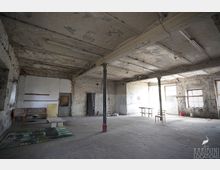 Großer, leerer Industrie- oder Lagerraum mit bröckelnder Betondecke, freiliegenden Trägern und Stützsäulen sowie abgenutzten, weiß gestrichenen Ziegelwänden. An der rechten Seite fallen Fensterlicht und einzelne verstreute Möbelstücke und Gegenstände in dem staubigen Raum auf.