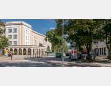 Straßenszene mit einem mehrstöckigen, beigefarbenen Gebäude im Hintergrund, das Arkadengänge im Erdgeschoss hat. Im Vordergrund stehen Bäume, geparkte Autos, eine öffentliche Uhr und ein Straßenschild "Poststraße".