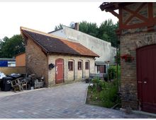 Innenhof mit gepflasterter Fläche vor einem kleinen Backsteingebäude mit Satteldach aus roten Ziegeln, roten Holztüren und mehreren kleinen Fenstern. Rechts ist der Rand eines weiteren Backsteinbaus mit Fachwerkdetails und roter Tür zu sehen, daneben ein schmaler Gartenstreifen mit Pflanzen.