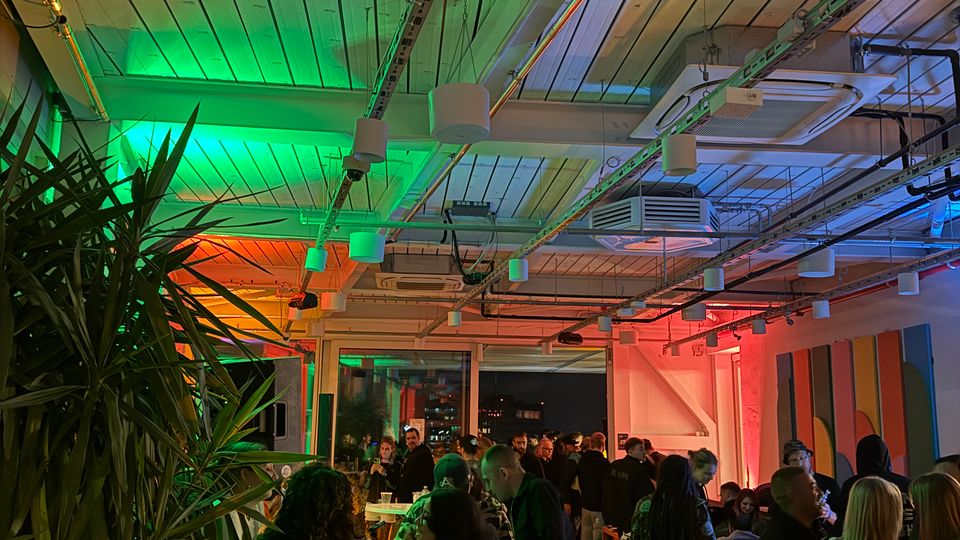 Interior of a modern event space with an exposed white-beam ceiling, visible ducts and cable trays, and colorful green, orange, and blue lighting. The room has tall plants, bar-height tables and stools, and large windows along one wall.