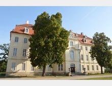 Historisches Gebäude mit cremefarbener Fassade, hohen Fenstern und einem roten Mansarddach, flankiert von großen Bäumen im Vordergrund. Im unteren Bereich führt eine Treppe zum Eingang mit einem verzierten Rundbogen.