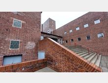 Backsteinarchitektur mit moderner Formsprache: Ein Gebäudekomplex aus rotem Klinkerstein mit Fenstern in unterschiedlichen Größen. Es sind mehrere Treppenaufgänge und eine erhöhte Eingangstür mit der Aufschrift „Stadtraum“ erkennbar, umgeben von klaren geometrischen Linien.