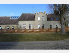 Großes zweistöckiges Wohnhaus mit grauer Fassade, Satteldach und weißen Fenstern, umgeben von einem Zaun aus Backsteinpfosten und Holzgittern. Im Vordergrund befindet sich eine gepflasterte Straße und ein Grünstreifen mit einem Baum.