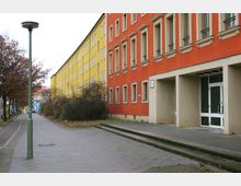 Straßenansicht mit zwei langen Wohngebäuden in Gelb und Rot, flankiert von einem Gehweg und kahlen Bäumen. Die Gebäude haben regelmäßig angeordnete Fenster und ein Eingangsportal mit einer Glastür.