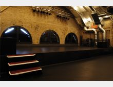 Innenraum eines Veranstaltungsraums mit Backsteinmauern, runden Fenstern und einem erhöhten Podest mit roten Stufen. An der Decke befinden sich metallene Lüftungsrohre sowie eine Beleuchtungsinstallation.