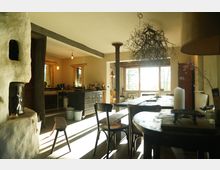 Wohnraum mit rustikalem Design, einschließlich Holzdetails und Lehmwänden. Im Vordergrund ein großer Holztisch mit Stühlen, im Hintergrund ein gemauerter Kaminofen, eine Theke und ein großes Fenster mit Blick ins Freie.