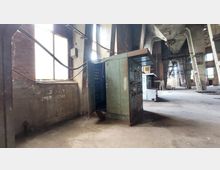 Innenansicht einer verlassenen Industriehalle mit hohen Betonsäulen und großen Fenstern, durch die Licht einfällt. Im Vordergrund steht ein veralteter, metallener Schaltschrank mit sichtbaren Gebrauchsspuren und Kabeln, der teilweise geöffnet ist.